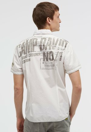 Junger Mann trägt ein weißes Kurzarmhemd mit großem verblasstem Schriftzug "CAMP DAVID NO 7" und weiteren kleineren gedruckten Details auf dem Rücken.