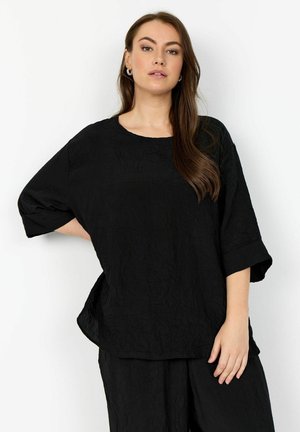 Donna con lunghi capelli castani che indossa una maglia nera morbida e testurizzata e pantaloni abbinati su sfondo bianco.