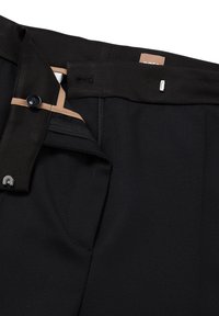 Černé kalhoty na míru s hladkou texturou, zipem na přední straně, zapínáním na knoflík a béžovým detailem v pase. Na látce je viditelný lineární vzor.