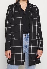 Manteau noir à manches longues avec un motif en grille blanche, porté sur un haut zippé noir et un jean taille haute bleu clair.