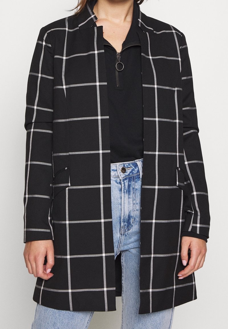 Manteau noir à manches longues avec un motif en grille blanche, porté sur un haut zippé noir et un jean taille haute bleu clair.