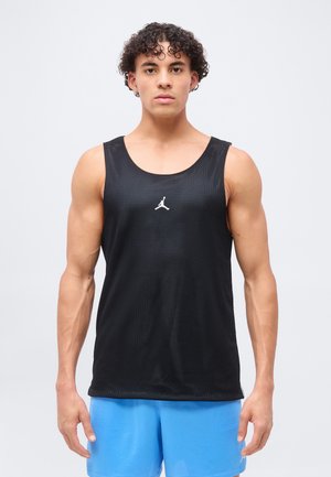 Giovane uomo con capelli ricci che indossa una canottiera sportiva nera senza maniche in rete e pantaloncini sportivi blu brillanti, in piedi davanti a uno sfondo semplice.