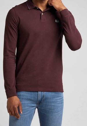 Mies, jolla on päällään pitkähihainen maroonin värinen Lee-logolla varustettu poolopaita ja vaaleansiniset farkut, seisoo yksinkertaista taustaa vasten.