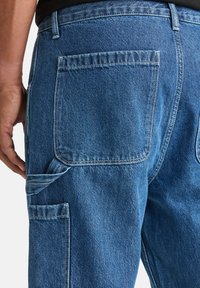 Jeans w średnim odcieniu niebieskiego z teksturowanym wykończeniem. Posiadają dwie tylne kieszenie oraz dodatkową boczną kieszeń z warstwowym wzornictwem.