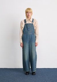 Ljusblå denimoveraller med avslappnad passform, som har sidofickor och justerbara axelremmar, i kombination med en beige och vit randig skjorta.