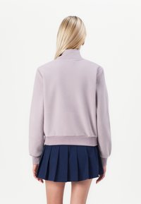Blonde person facing away wearing a light purple fleece jacket and a navy blue pleated skirt against a plain white background.