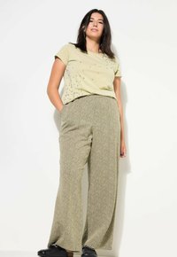 Olivově zelené tričko s dekorativními průhlednými korálky a volnými kalhotami s houndstooth vzorem. Modelka stojí s rukama v kapsách.