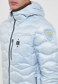 Giacca piumino azzurra chiara con design trapuntato, dotata di cappuccio, zip frontale e patch con logo sulla spalla. Finishing testurizzato e lucido.