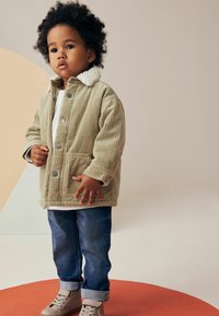 Giacca in corduroy beige con colletto in pelliccia bianca, chiusura frontale con bottoni, tasche laterali, abbinata a jeans blu e sneakers grigio chiaro.