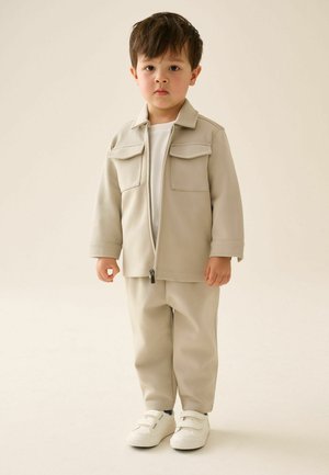 Junge steht und trägt eine beige Reißverschlussjacke, passende Hose, weißes Hemd und weiße Sneaker vor einem einfarbigen hellen Hintergrund.