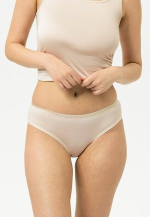 Frau trägt beige Tanktop und passende Unterwäsche, zieht den Saum des Tanktops mit beiden Händen gegen einen einfarbigen Hintergrund nach unten.