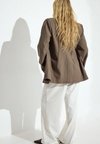 Brown tailored blazer with flared sleeves, paired with wide white trousers. Smooth texture, minimalist design, and a visible back seam.