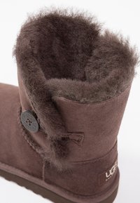 Brązowy zamszowy but z teksturowaną wewnętrzną podszewką, ozdobnym guzikiem i pętlą, zwężającym się kształtem oraz płaską podeszwą.