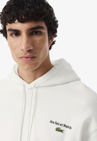 Joven con cabello rizado y oscuro que lleva una sudadera blanca con el texto "Jeu Set et Match" y un logo de cocodrilo verde en el pecho.