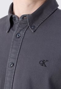 Harmaa nappipaita teksturoidulla kankaalla, jossa on kaulus, mustat napit ja rinnassa kirjailtu cK-logo.