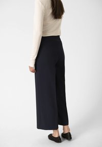Pantaloni a gamba larga blu navy con una texture liscia, vita alta e dettagli sulle cuciture, abbinati a una blusa leggera a maniche lunghe e scarpe flat.