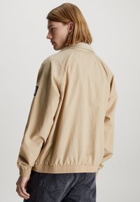Chaqueta bomber beige con un ajuste relajado, puños y dobladillo elásticos, y un pequeño parche negro con la marca en la manga izquierda. Tejido de algodón.