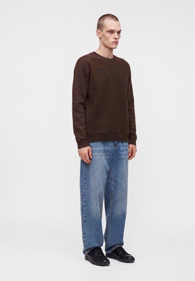 Jeune homme debout, portant un sweat-shirt marron foncé, un jean bleu ample et des baskets noires sur un fond clair uni.