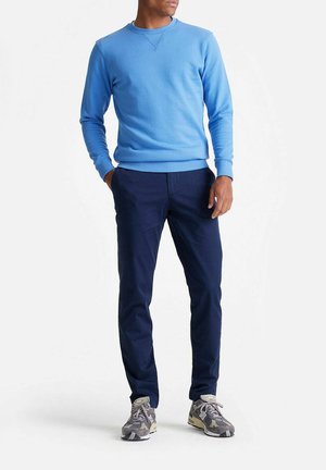 Mann trägt hellblauen Sweatshirt, dunkelblaue Hose und graue Sportschuhe, steht mit einer Hand in der Tasche vor weißem Hintergrund.