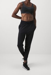 Schwarzer gerippter Sport-BH kombiniert mit schwarzen Jogginghosen mit elastischem Bund und Zugband. Das Model trägt schwarze Sportschuhe.