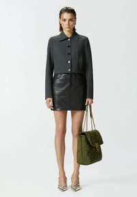 Giacca grigia alla cintura con bottoni neri, abbinata a una gonna in pelle nera. Tiene in mano una borsa trapuntata verde con tracolle dorate. Tacchi argento.