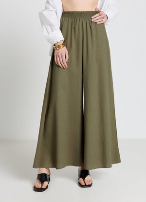 Frau trägt hoch taillierte, weit geschnittene olivgrüne Hosen, schwarze Sandalen mit offenen Zehen, weiße Blusenärmel sowie goldene Armbänder und Ringe.