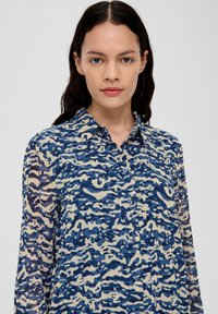 Chemise à manches longues bleu et beige avec un motif ondulé, fermeture à boutons à l'avant, col et texture de tissu douce.
