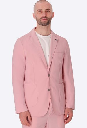 Uomo con barba che indossa una giacca da abito rosa chiaro sopra una camicia bianca, in piedi davanti a uno sfondo bianco uniforme.