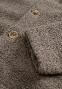 Grauer Strukturstoff mit einer flauschigen Oberfläche, versehen mit zwei runden, transparenten Knöpfen. Der Stoff ist gefaltet und zeigt seine Dicke und Weichheit.