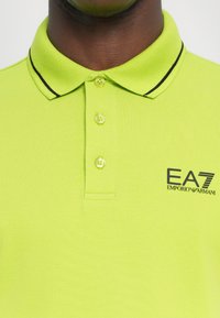 Limonkovo zelená polo košeľa vyrobená z textúrovanej látky má golier s čiernymi prúžkami, tri gombíky a logo EA7 Emporio Armani na hrudi.