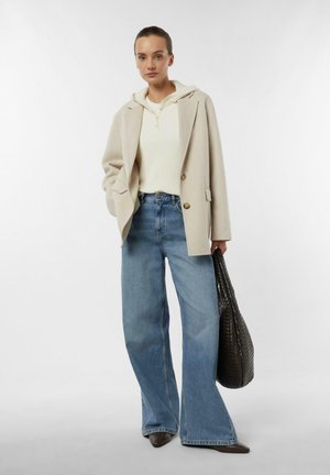 Kobieta w beżowej marynarce na białej bluzie z kapturem i prążkowanym swetrze, szerokie niebieskie dżinsy, trzyma dużą czarną plecioną torbę.