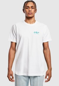 Camiseta de algodón blanca con un logo turquesa en el lado izquierdo del pecho. Manga corta, corte clásico y textura suave. Combinada con jeans de color azul claro.