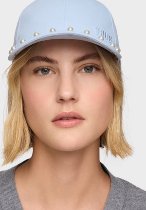 Mujer con cabello rubio corto que lleva una gorra azul claro decorada con perlas y un suéter gris, expresión facial neutra.