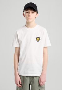 T-shirt bianca in cotone con un design di fiore giallo e testo nero sulla parte frontale, abbinata a pantaloni corti verde oliva e un cappellino nero.