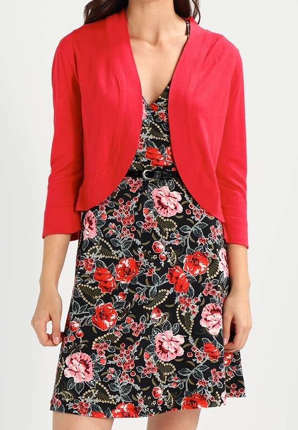 Femme portant une robe noire à fleurs rouges et roses, associée à un cardigan court rouge vif à manches trois-quarts.