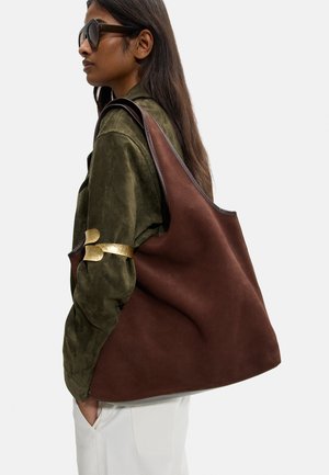 Kobieta w okularach przeciwsłonecznych, ubrana w zieloną kurtkę i białe spodnie, niosąca dużą brązową skórzaną torbę na ramieniu na szarym tle.