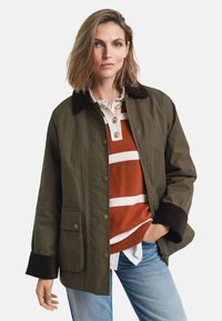 Olive green jacket with a brown collar, large front pockets, and a relaxed fit. Layered over an orange and white striped sweater.
