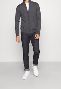 Grå zip-up tröja med ståkrage, bärs över en vit t-shirt, ihop med mörka jeans och vita sneakers. Mjuk textur, figursydd design.