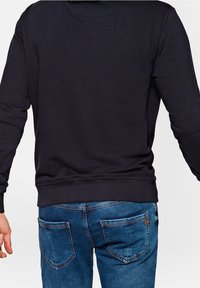 Černá mikina s kulatým výstřihem, charakterizovaná dlouhými rukávy a jednobarevným designem. V kombinaci s modrými džíny, které mají dvě zadní kapsy.