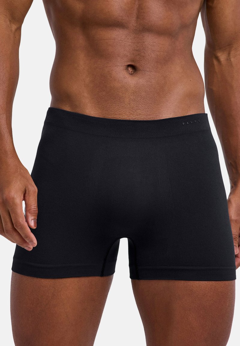 Torso maschile che indossa boxer aderenti neri, in piedi con le braccia rilassate lungo i fianchi contro uno sfondo semplice.