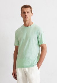 Helles mintgrünes T-Shirt aus einem weichen, strukturierten Stoff; mit Rundhalsausschnitt und kurzen Ärmeln, getragen mit hellen Hosen.