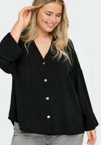 Črna srajca z gumbki, z V-izrezom, dolgimi rokavi in srebrnimi gumbi; izdelana iz teksturirane tkanine, sproščenega kroja in ukrivljenega roba.