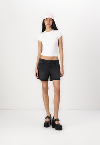 Chemise ajustée à manches courtes blanche, shorts en denim noir, et chaussures à plateforme noires avec des brides à la cheville. Casquette rose avec du texte sur le devant.
