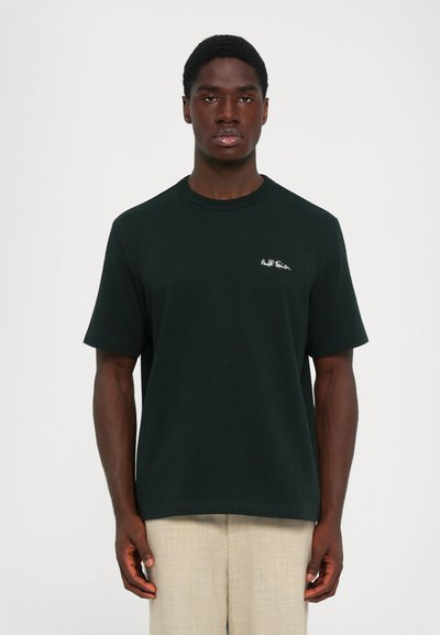 Mand iført en sort Paul Smith t-shirt og beige bukser, stående mod en ensfarvet lys baggrund, vendt fremad med afslappede arme.