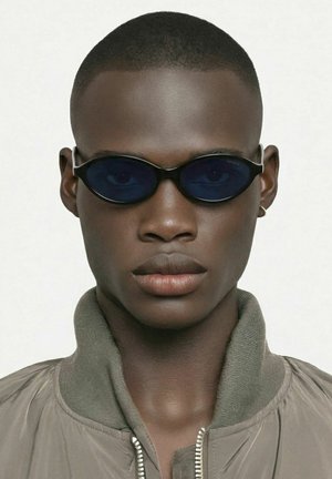 Jeune homme aux cheveux coupés court, portant de petites lunettes de soleil ovales foncées, une veste grise zippée et une petite boucle d'oreille en anneau sur fond blanc.