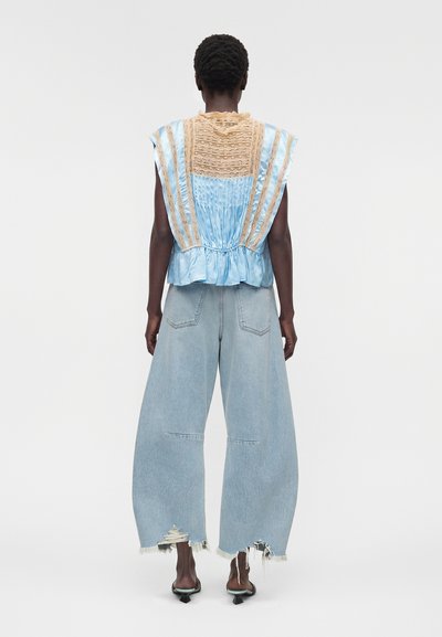 Άτομο που φοράει μπλε αμάνικη μπλούζα με μπεζ δαντελένιες λεπτομέρειες και φαρδιά ανοιχτό μπλε τζιν με ξεφτισμένα τελειώματα, στέκεται μπροστά σε απλό λευκό φόντο.