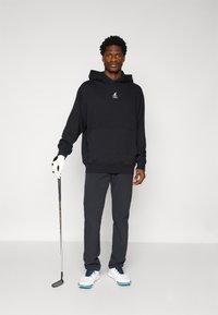 Sweatshirt preto com um bolso frontal e logótipo. Calças cinzentas, sapatos de golfe brancos com detalhes azuis. Pessoa segurando um taco de golfe, vestindo uma luva branca.