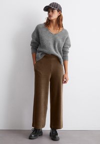 Eine Frau, die einen grauen Strickpullover, braune weit geschnittene Hosen, schwarze Schnürschuhe und eine karierte Schiebermütze trägt, steht vor einer weißen Wand.