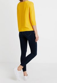 Gelber Strickpullover mit Dreiviertelärmeln, kombiniert mit dunkelblauen Skinny-Jeans und weißen Sneakers. Rückansicht, vor einem schlichten Hintergrund.