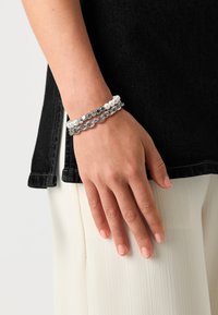 Bracelet en chaîne argentée avec des perles blanches et des perles métalliques ; présente un design superposé avec des textures et des formes variées sur un poignet.